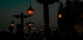 Several hanging lanterns emit a warm glow in an outdoor restaurant setting, with silhouettes of people and wooden structures. The sky is dark, adding a sense of evening ambiance.
