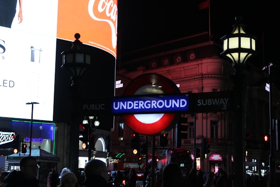 Nighttime scene featuring a bright illuminated London Underground sign amidst a busy urban environment, with various advertisements and architectural elements visible in the background.