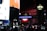 Nighttime scene featuring a bright illuminated London Underground sign amidst a busy urban environment, with various advertisements and architectural elements visible in the background.