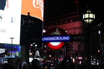 Nighttime scene featuring a bright illuminated London Underground sign amidst a busy urban environment, with various advertisements and architectural elements visible in the background.
