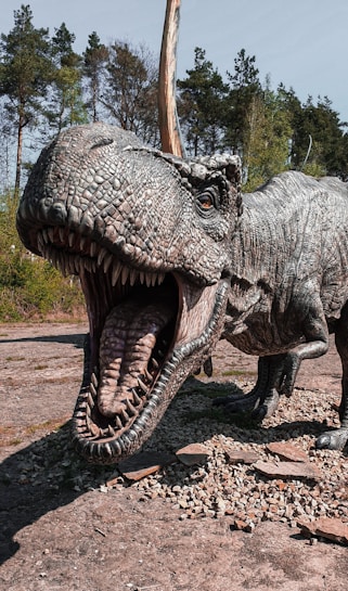 A large and detailed dinosaur sculpture with an open mouth and sharp teeth is positioned in an outdoor setting. The dinosaur's skin texture appears scaly and realistic, with a predominantly gray color. In the background, there are tall, dense trees under a clear sky, adding a natural setting to the scene.