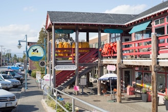 A street scene featuring a wooden building with multiple signs, including one for Vancouver Whale Watch and another for Mini Donuts. Brightly colored jackets or coats hang on the balcony railing. Several people are walking along the sidewalk near parked cars. An umbrella shades an outdoor seating area near smaller signs.