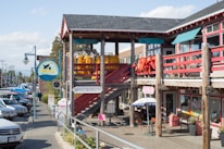 A street scene featuring a wooden building with multiple signs, including one for Vancouver Whale Watch and another for Mini Donuts. Brightly colored jackets or coats hang on the balcony railing. Several people are walking along the sidewalk near parked cars. An umbrella shades an outdoor seating area near smaller signs.