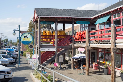 A street scene featuring a wooden building with multiple signs, including one for Vancouver Whale Watch and another for Mini Donuts. Brightly colored jackets or coats hang on the balcony railing. Several people are walking along the sidewalk near parked cars. An umbrella shades an outdoor seating area near smaller signs.