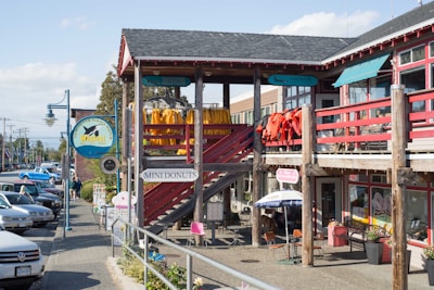 A street scene featuring a wooden building with multiple signs, including one for Vancouver Whale Watch and another for Mini Donuts. Brightly colored jackets or coats hang on the balcony railing. Several people are walking along the sidewalk near parked cars. An umbrella shades an outdoor seating area near smaller signs.