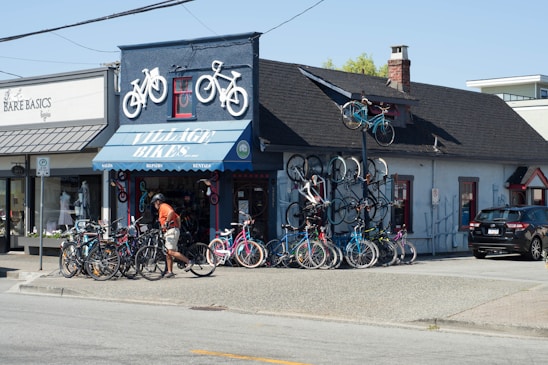 A bicycle shop with several bikes displayed outside and two on the roof as decorative elements. A person in an orange shirt and shorts is walking near the bikes. The building has a blue awning with the text 'VILLAGE BIKES' and a smaller adjacent shop named 'BARE BASICS.' There is a parking area with a black car on the right.