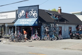 A bicycle shop with several bikes displayed outside and two on the roof as decorative elements. A person in an orange shirt and shorts is walking near the bikes. The building has a blue awning with the text 'VILLAGE BIKES' and a smaller adjacent shop named 'BARE BASICS.' There is a parking area with a black car on the right.