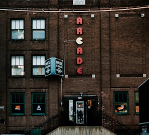 A brick building facade featuring a vertical neon sign spelling 'ARCADE' with a Pac-Man symbol. Several windows are present, some displaying arcade and gaming logos like Midway and Atari. The entrance has a vintage aesthetic with multiple posters on the door. The building has a nostalgic charm, hinting at retro gaming culture.