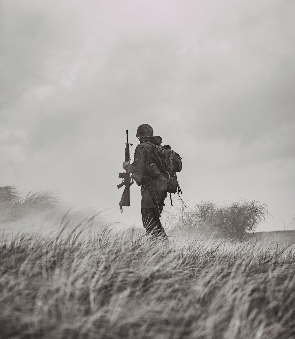 Foto en escala de grises de un hombre con chaqueta negra y pantalones sosteniendo un rifle