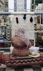 A person sits in front of a loom, weaving a textile piece with various colors of yarn hanging above. The background features an array of intricately patterned carpets displayed on a wall. The individual is wearing a patterned headscarf and a pink garment, seated on a bench covered with a decorative textile.