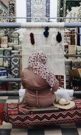 A person sits in front of a loom, weaving a textile piece with various colors of yarn hanging above. The background features an array of intricately patterned carpets displayed on a wall. The individual is wearing a patterned headscarf and a pink garment, seated on a bench covered with a decorative textile.
