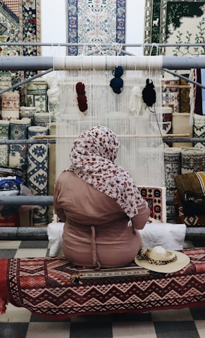 A person sits in front of a loom, weaving a textile piece with various colors of yarn hanging above. The background features an array of intricately patterned carpets displayed on a wall. The individual is wearing a patterned headscarf and a pink garment, seated on a bench covered with a decorative textile.