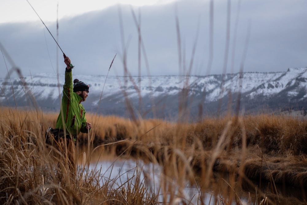 man in green jacket and black pants holding fishing rod standing on brown grass field during