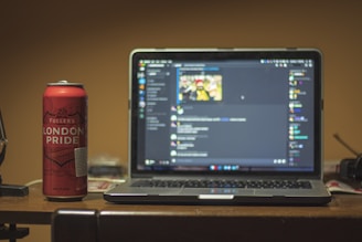 A can of Fuller's London Pride is positioned next to an open laptop displaying a dark-themed chat application screen. The scene is set on a wooden table with a few indistinct items in the background, suggesting a casual or home environment.
