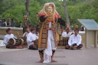 A person dressed in traditional Balinese attire is performing a dance in front of a group of musicians. The dancer wears a colorful costume with intricate patterns and a white mask. The musicians, sitting cross-legged, are playing traditional instruments while dressed in white shirts and matching headgear. The background consists of lush greenery and a stone path, creating an outdoor setting.