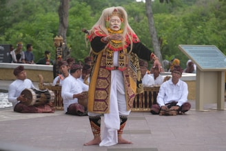 A person dressed in traditional Balinese attire is performing a dance in front of a group of musicians. The dancer wears a colorful costume with intricate patterns and a white mask. The musicians, sitting cross-legged, are playing traditional instruments while dressed in white shirts and matching headgear. The background consists of lush greenery and a stone path, creating an outdoor setting.