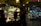 A dimly lit flower shop at night with a variety of colorful flowers on display. There are people walking by and one person standing inside the shop, slightly obscured by shadows. The wet pavement reflects the street lights, adding a moody ambiance.