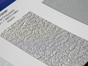A close-up of a textured silver surface with a shimmering, metallic finish. The surface is displayed on a sample card with printed text on the left featuring technical details.
