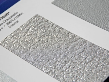 A close-up of a textured silver surface with a shimmering, metallic finish. The surface is displayed on a sample card with printed text on the left featuring technical details.