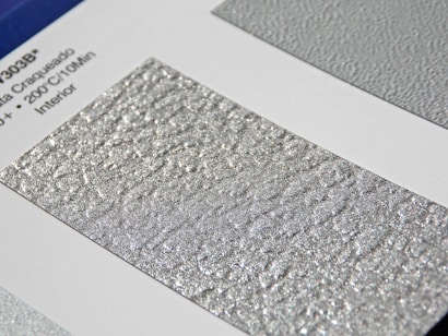 A close-up of a textured silver surface with a shimmering, metallic finish. The surface is displayed on a sample card with printed text on the left featuring technical details.