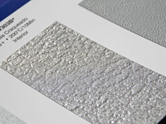 A close-up of a textured silver surface with a shimmering, metallic finish. The surface is displayed on a sample card with printed text on the left featuring technical details.