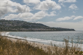 A coastal scene featuring a sandy beach with people strolling along the shoreline. The ocean glistens under a partly cloudy sky, while a hillside community with numerous houses stretches across the horizon.