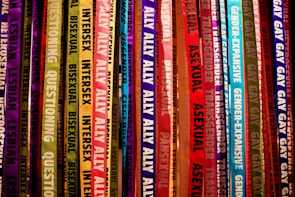 Colorful ribbons featuring words related to gender identity and sexual orientation hang vertically. Each ribbon displays labels like lesbian, bisexual, intersex, transgender, and ally, among others. The ribbons are in a vibrant array of shades, creating a rainbow effect.