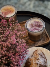 Two glasses of creamy, frothy coffee are adorned with vibrant sprinkles and sit on a wooden tray. Beside them, a cluster of delicate pink flowers adds a touch of elegance, and a croissant dusted with powdered sugar rests on a white plate.