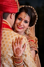 A bride adorned in traditional Indian attire, featuring detailed jewelry and henna designs while embracing her partner. The bride is smiling, with decorative elements including a bindi, earrings, and an intricate hairstyle.