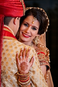 A bride adorned in traditional Indian attire, featuring detailed jewelry and henna designs while embracing her partner. The bride is smiling, with decorative elements including a bindi, earrings, and an intricate hairstyle.