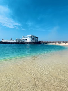 A serene beach scene with golden sand leading into clear turquoise waters. In the background, there is a large, sophisticated building by the water and a wooden pier extending into the sea. The sky is bright and clear, adding to the tranquil atmosphere.