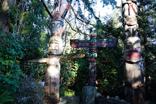 Three intricately carved totem poles stand amidst lush green foliage. Each pole features vivid colors and traditional designs, with faces and animal motifs. The central pole has a wide wingspan, suggesting a bird-like creature.