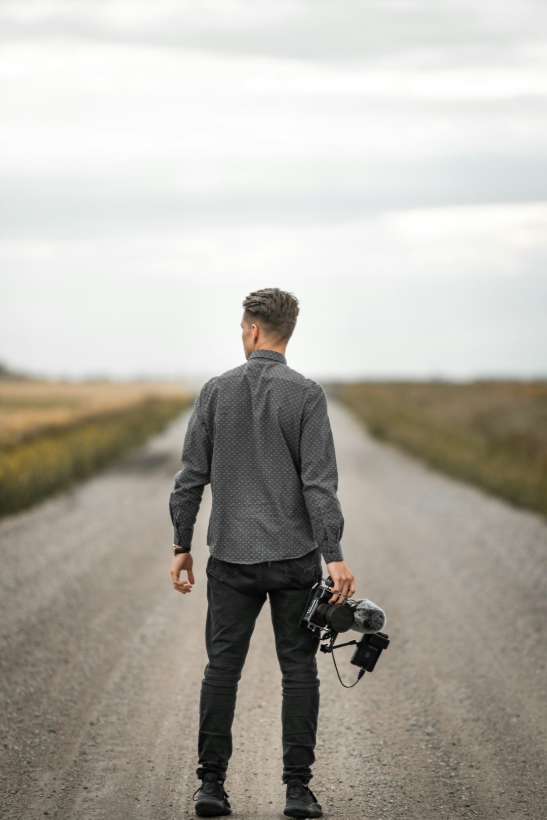 man in gray sweater and black pants holding black dslr camera standing on gray asphalt road