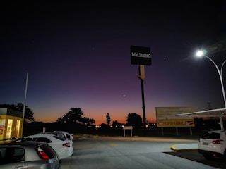 A parking lot with several cars is illuminated by a streetlight. The sky is a gradient of deep blue to orange, indicating a sunset or dusk. A prominent sign reading 'Madero' is visible, along with another sign advertising a factory outlet. Trees and other vegetation can be seen in the background silhouetted against the sky.