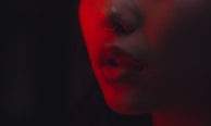 A close-up shot of a person's lower face, focusing on their nose and slightly parted lips. The lighting is dramatic, with a strong red hue casting shadows and enhancing features like the nose ring.