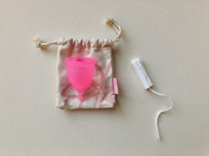 A bright pink menstrual cup is placed on top of a small, patterned drawstring pouch with a rope closure. Nearby, there is a white tampon with an extended string.