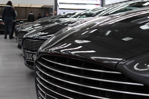 A row of luxury cars is parked indoors, showcasing their sleek design and glossy finishes. The cars are aligned closely, with a focus on their distinctive front grilles and hoods. The setting appears to be a showroom with reflective lighting casting highlights on the car surfaces. Several people are visible in the background, likely viewing or interacting with the cars.
