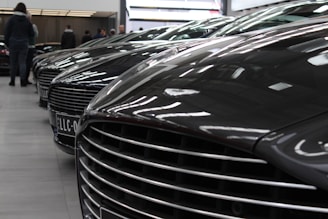A row of luxury cars is parked indoors, showcasing their sleek design and glossy finishes. The cars are aligned closely, with a focus on their distinctive front grilles and hoods. The setting appears to be a showroom with reflective lighting casting highlights on the car surfaces. Several people are visible in the background, likely viewing or interacting with the cars.