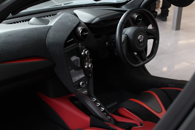 The image features the interior of a luxury car, showcasing a sleek and modern design. The dashboard and steering wheel are crafted with fine materials, predominantly in black, with red accents on the seats. The center console houses a sophisticated set of controls and a display. The steering wheel has a badge at its center, and the overall ambiance is of high-end automotive craftsmanship.