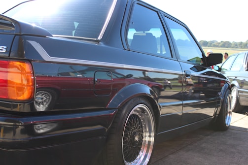 A vintage car is parked outdoors, showcasing its glossy black finish and polished rims under bright sunlight. The car's rear section, including the taillight and rear window, is prominently focused. Reflection on the car's surface indicates other vehicles and elements of the surrounding environment.