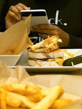 A person holds a smartphone while seated at a table with a plate of food. The food consists of a sandwich with thick slices of bread and a pickle on the side. A portion of fries is visible in the foreground, suggesting a casual dining setting.