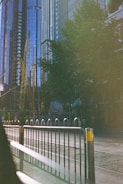 Tall skyscrapers with reflective glass windows rise above the city, interspersed with construction cranes. A roadway runs alongside a metal barrier, and a tram is visible in the street. Green trees provide some foliage amidst the urban landscape.