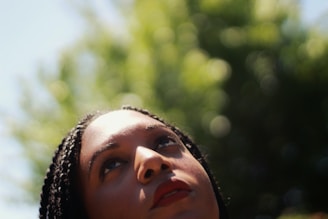 A close-up of a person looking upwards with a thoughtful expression. The background consists of lush greenery out of focus, suggestive of a natural setting.