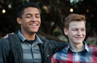 Two young people are standing close to each other with smiles on their faces. One appears to have his arm around the other’s shoulder, suggesting camaraderie and friendship. They are outdoors, with blurred foliage and lights in the background.