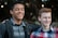 Two young people are standing close to each other with smiles on their faces. One appears to have his arm around the other’s shoulder, suggesting camaraderie and friendship. They are outdoors, with blurred foliage and lights in the background.