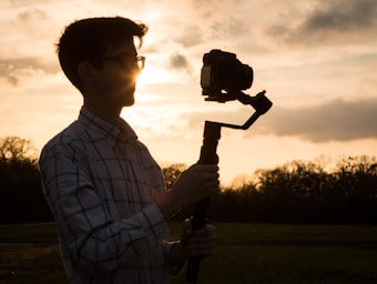 A silhouette of a person holding a camera on a gimbal against a sunset background. The sun is shining brightly, partially obscured by the person's head, creating a dramatic outline. The landscape features a field with trees in the distance, under a sky with scattered clouds.