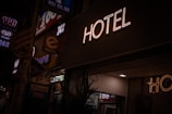 A neon sign reads 'HOTEL' illuminated against a dark background, with dim street signage nearby indicating a 24-hour convenience store. A faint indoor light spills onto the scene, partially lighting up the surroundings.