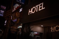 A neon sign reads 'HOTEL' illuminated against a dark background, with dim street signage nearby indicating a 24-hour convenience store. A faint indoor light spills onto the scene, partially lighting up the surroundings.