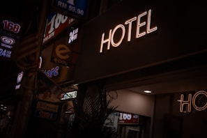 A neon sign reads 'HOTEL' illuminated against a dark background, with dim street signage nearby indicating a 24-hour convenience store. A faint indoor light spills onto the scene, partially lighting up the surroundings.