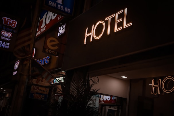 A neon sign reads 'HOTEL' illuminated against a dark background, with dim street signage nearby indicating a 24-hour convenience store. A faint indoor light spills onto the scene, partially lighting up the surroundings.